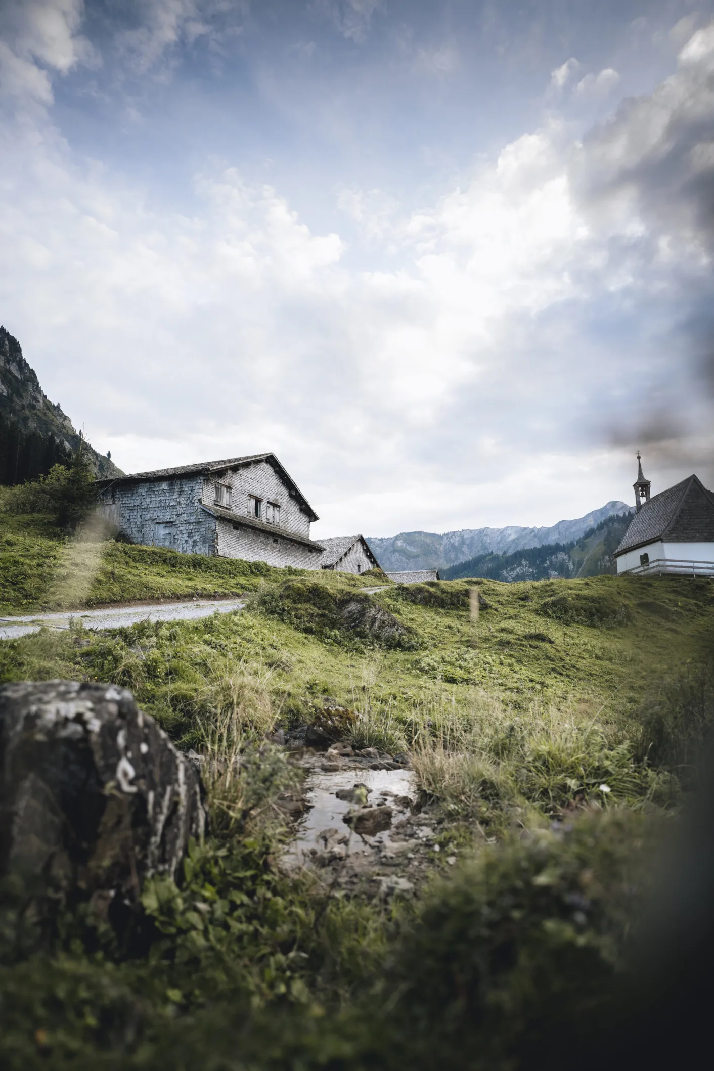 Traditionelle Alphütten und eine kleine Kapelle aus Stein auf grünen Hügeln vor einer beeindruckenden Bergkulisse unter bewölktem Himmel, die das authentische und idyllische Ambiente der alpinen Umgebung nahe dem Aparthotel Hubertus in Mellau einfängt.